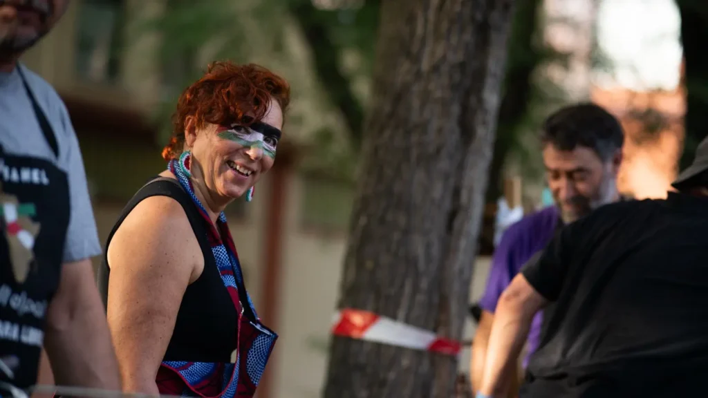 Una persona con el pelo corto y rizado de color rojo y la cara pintada de colores sonríe mientras está de pie al aire libre cerca de otras personas, con árboles y cinta roja y blanca visibles en el fondo.