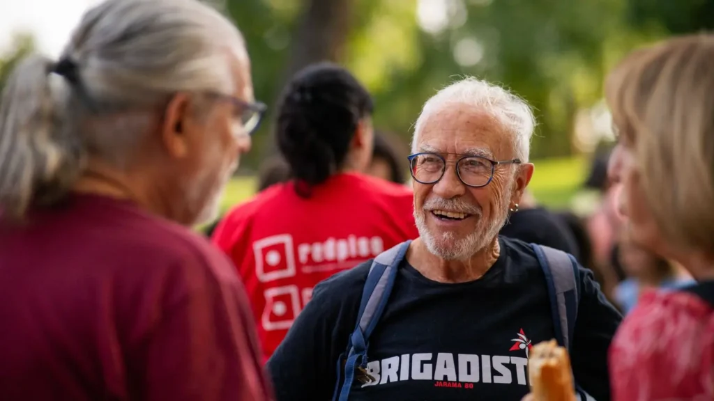 Un hombre mayor con gafas y barba canosa sonríe mientras habla con dos personas al aire libre. Lleva una camiseta negra de 