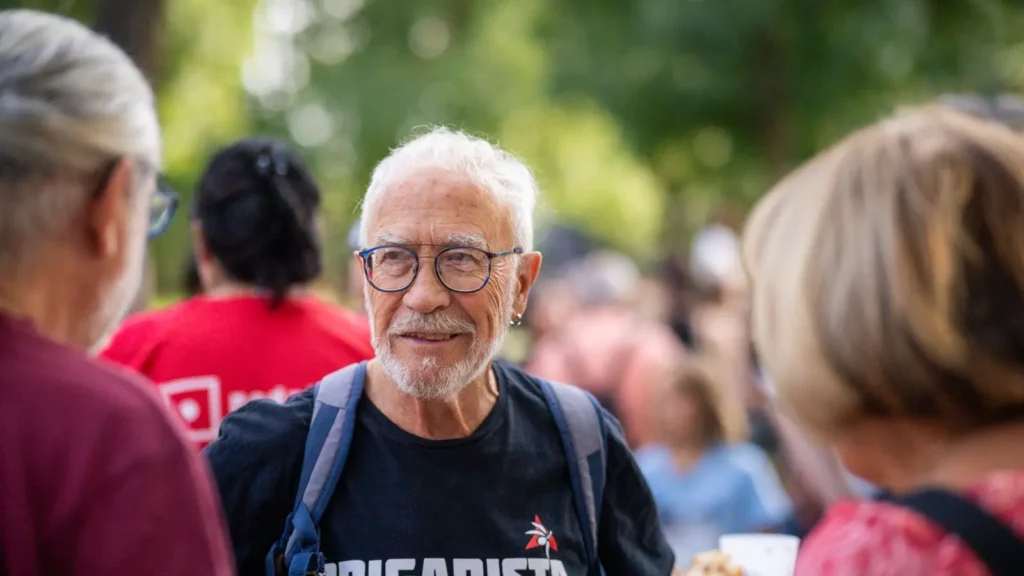 Un hombre mayor con pelo canoso, barba y gafas lleva una camiseta negra y una mochila, de pie al aire libre entre un grupo de personas. Al fondo se ven árboles y vegetación, donde se celebran las Fiestas Solidarias de La Pablo 2025.
