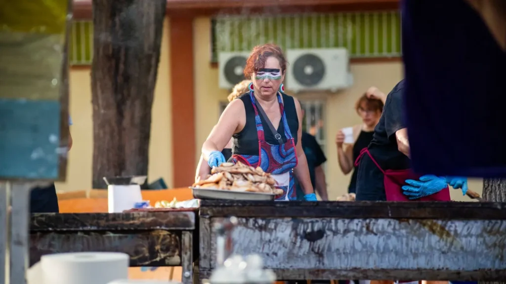 Una mujer con guantes y gafas protectoras se encuentra en una parrilla al aire libre, sosteniendo una bandeja de comida cocinada para disfrutarla en las Fiestas Solidarias de La Pablo 2025.