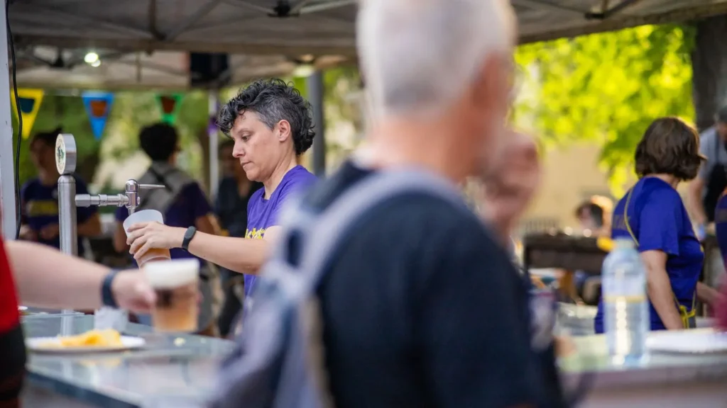 Una persona con camisa morada sirve una bebida de un grifo en las Fiestas Solidarias de La Pablo 2025, con varias personas en primer plano y al fondo. Detrás de la multitud se ven coloridos banderines y árboles.