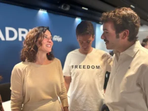 Tres personas conversan en un interior. La mujer de la izquierda sonríe con un top beige. El hombre del centro, con una camiseta blanca de LIBERTAD, es Jesús Martínez Caballero, de NNGG Madrid. El hombre de la derecha lleva una camiseta clara.