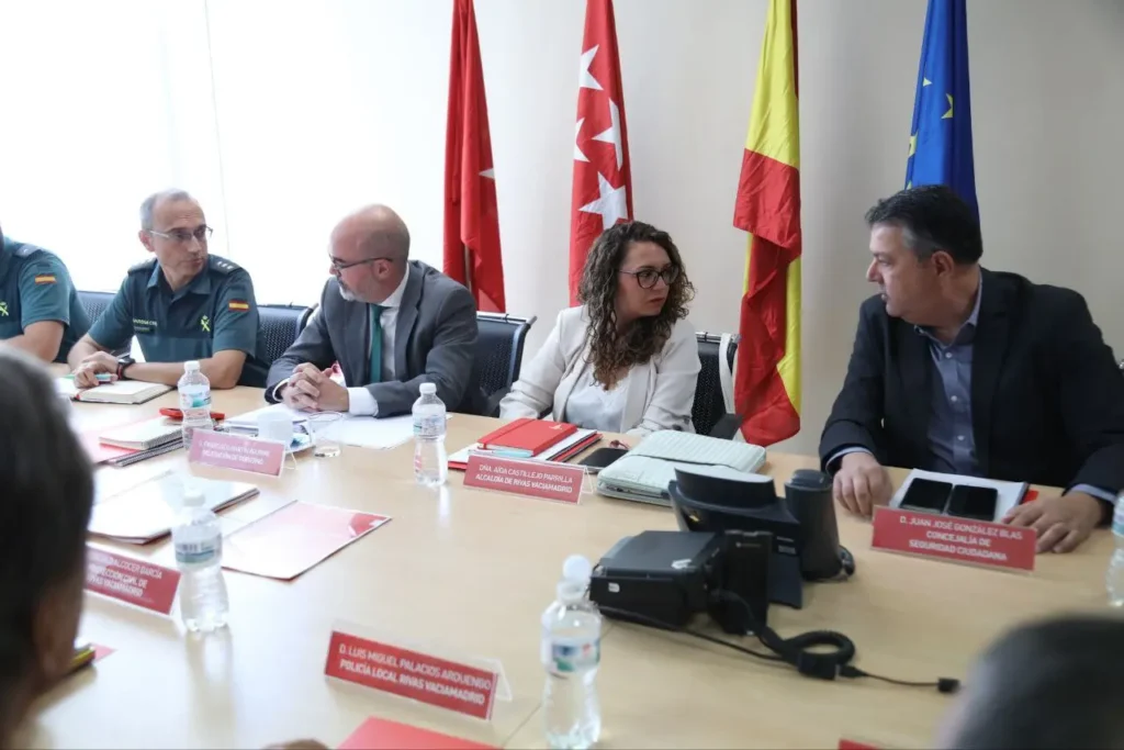 Varias personas se sientan en una mesa de reuniones con documentos y botellas de agua, discutiendo temas como robos en establecimientos. Tres banderas -Madrid, España y la Unión Europea- se yerguen al fondo, con placas de identificación y carpetas visibles ante los asistentes.