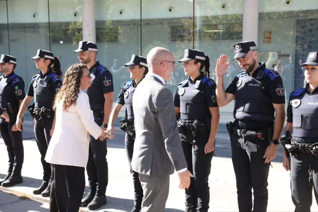 Un grupo de policías uniformados en fila al aire libre. Dos personas en traje de negocios saludan a los agentes; un agente levanta la mano en un gesto. La escena, posiblemente en Rivas, tiene ventanas de cristal y una zona iluminada por el sol al fondo.