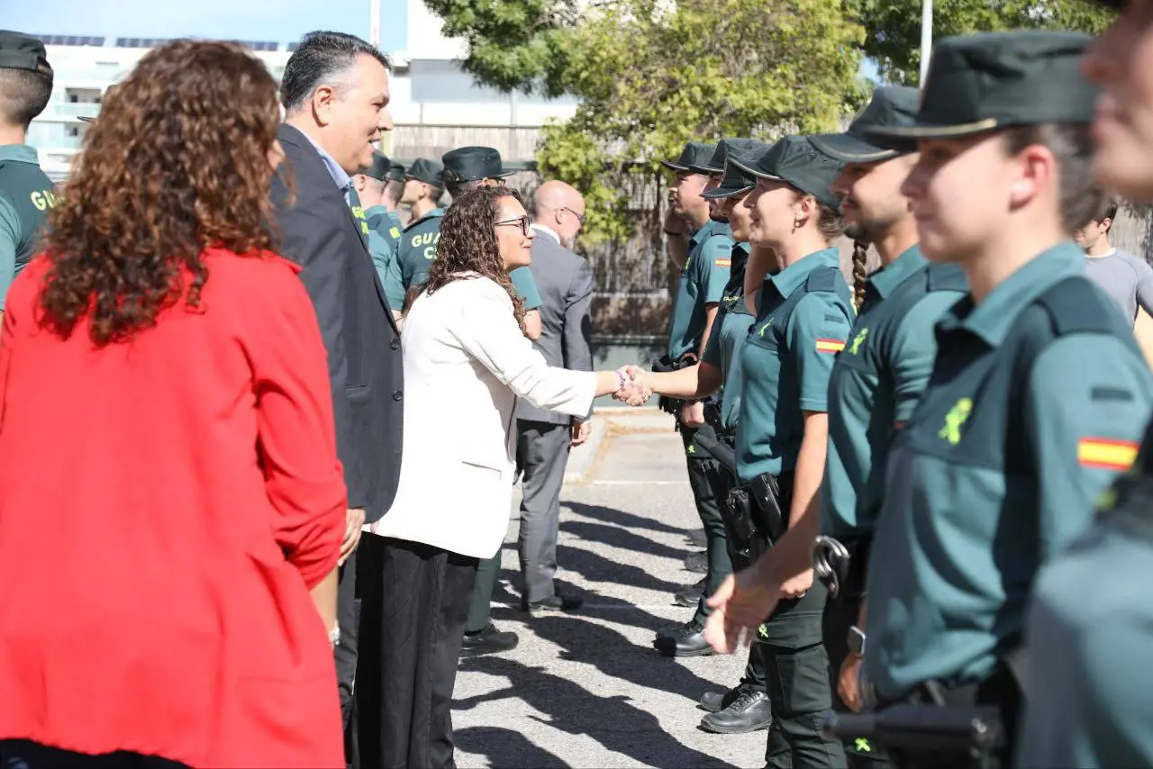 Una mujer con americana blanca estrecha la mano de un agente uniformado al aire libre, posiblemente tras una reunión informativa sobre robos en establecimientos. Varias personas, entre ellas un hombre trajeado y otras de paisano, también están presentes.