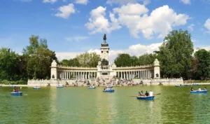 La gente rema en barcas azules en un gran lago de El Retiro, frente a un gran monumento de piedra con columnas y una estatua en el centro, rodeado de árboles y un cielo parcialmente nublado, que ofrece accesibilidad a todos los visitantes.