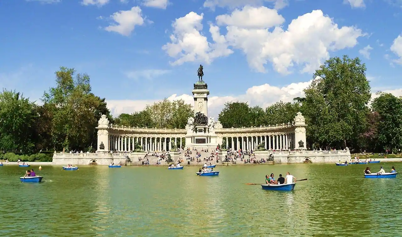 La gente rema en barcas azules en un gran lago de El Retiro, frente a un gran monumento de piedra con columnas y una estatua en el centro, rodeado de árboles y un cielo parcialmente nublado, que ofrece accesibilidad a todos los visitantes.