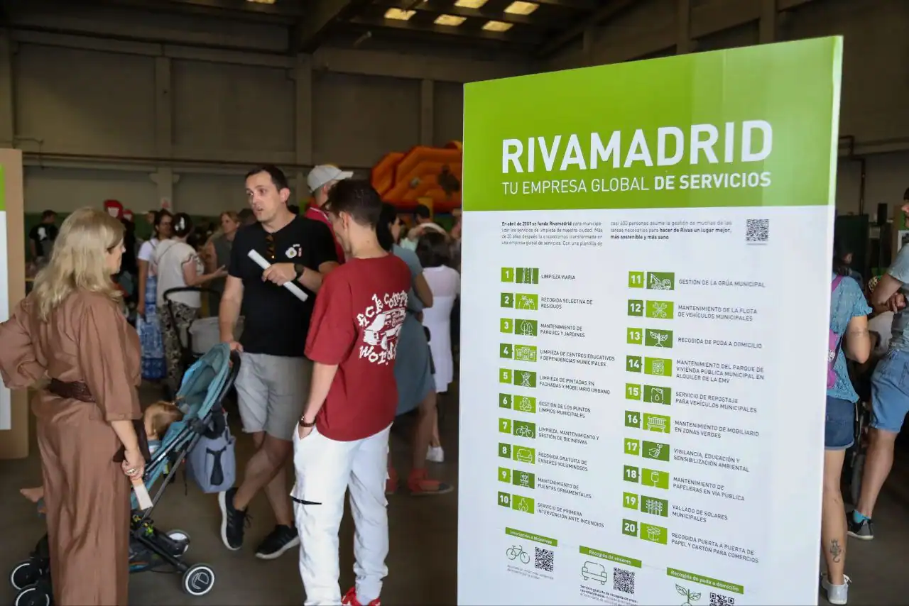 Jornada Puertas Abiertas Rivamadrid 2025 La gente se para y conversa en el interior cerca de un gran cartel verde y blanco de Rivamadrid en el que se lee "TU EMPRESA GLOBAL DE SERVICIOS." La zona, posiblemente parte de un evento de bolsa de empleo, está abarrotada de gente con una atracción hinchable al fondo.