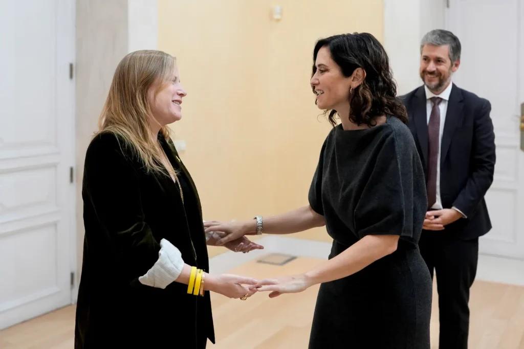 Dos mujeres se dan la mano y se sonríen en una habitación muy iluminada, mientras el consejero Miguel Ángel García, de pie en el fondo, las observa y sonríe.