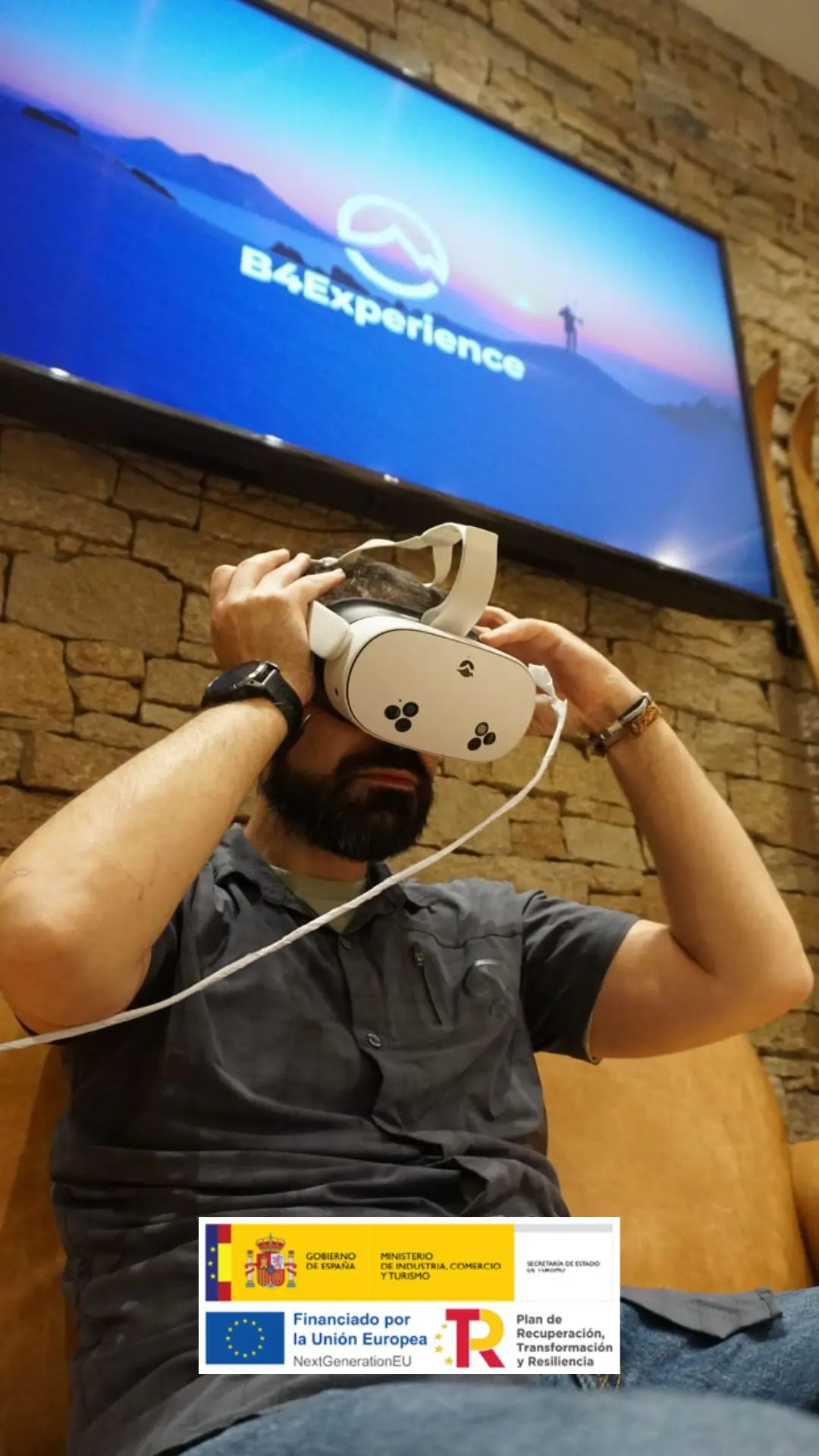 Un hombre con barba lleva unos auriculares de realidad virtual delante de un muro de piedra y una pantalla en la que aparece el logotipo de B4Experience, que explora actividades de aventura. En la parte inferior de la imagen aparecen los logotipos de financiación del Gobierno y de la UE.
