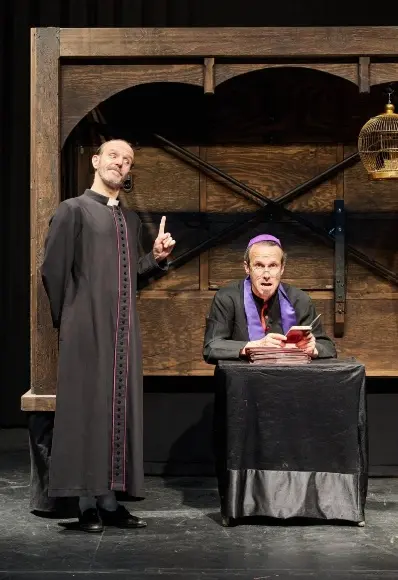 Dos hombres con trajes teatrales actúan en el escenario; uno gesticula con un dedo levantado, mientras que el otro, vestido con atuendo clerical y un casquete morado, lee en un libro rojo, aludiendo al tema de las inmatriculaciones de la Iglesia en Rivas.