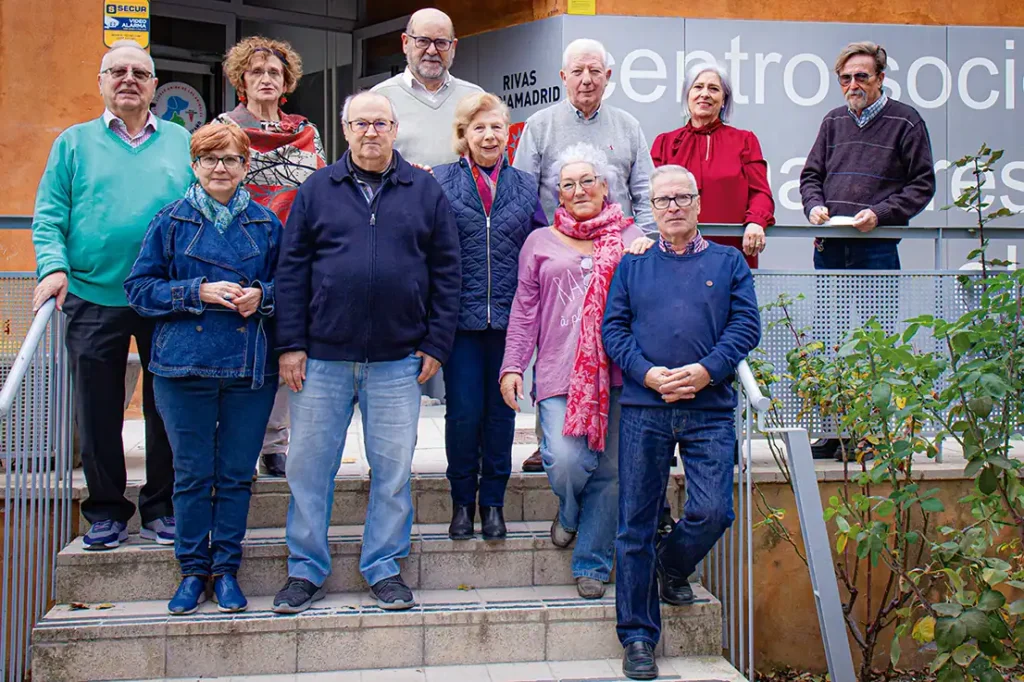 Consejo Rector de los centros de mayores de Rivas - AYUNTAMIENTO DE RIVAS. Se ve a un grupo de once mayores de Rivas, vestidas con ropa informal, permanecen juntas en unas escaleras al aire libre ante un edificio con un cartel en el que se lee parcialmente centro social. Algunos sonríen, otros cruzan los brazos.