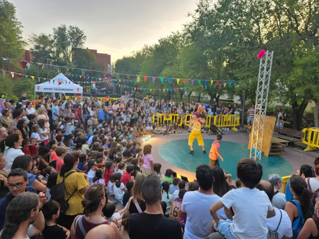 Una gran multitud se reúne en torno a un escenario circular al aire libre en las Fiestas Solidarias, observando a dos artistas, uno de ellos vestido de amarillo. Árboles, coloridos banderines y una carpa decoran el soleado escenario mientras La Pablo actúa en apoyo de Palestina.
