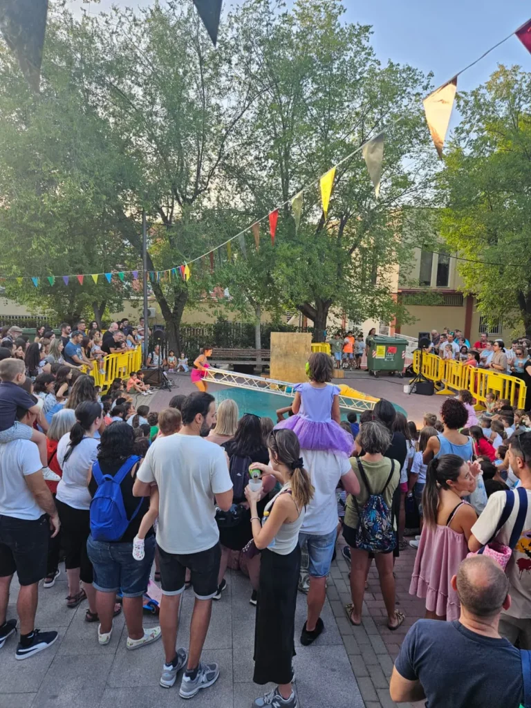 Una multitud de personasen las Fiestas Solidarias de La Pablo 2025, entre ellas niños y adultos, observa una actuación al aire libre en un parque durante las Fiestas solidarias organizadas por La Pablo. En lo alto cuelgan banderolas de colores y barricadas amarillas rodean la zona, con árboles y edificios al fondo.