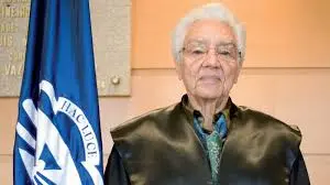 Una persona mayor, con pelo corto y blanco, gafas, toga académica oscura y corbata verde, junto a una bandera azul y blanca con un emblema que recuerda a la matemática española MARÍA WONENBURGER (1927-2014).
