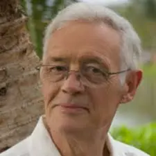 Un hombre mayor, con el pelo corto y canoso y gafas, está de pie al aire libre, cerca de un árbol, con una camisa blanca de cuello. El fondo verde, ligeramente difuminado, evoca la calma reflexiva que suele verse en los retratos de la matemática española MARÍA Wonenburger.