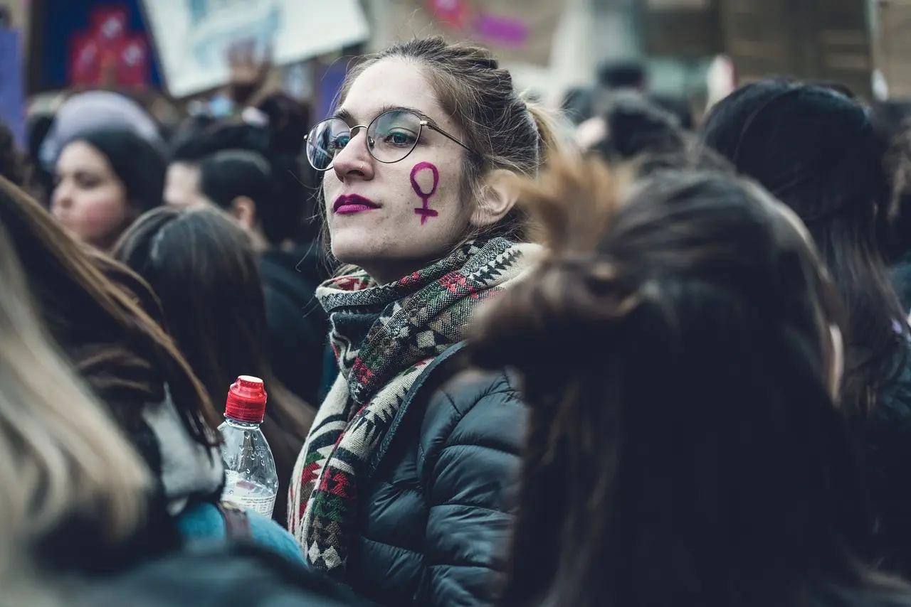 Mujeres que transforman. Una mujer con gafas y pañuelo sujeta una botella entre la multitud. Lleva un símbolo feminista morado dibujado en la mejilla, una poderosa imagen captada por Borrador automático durante una reunión pública o una protesta.