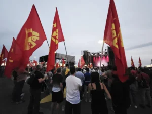 Una multitud se reúne al aire libre en Rivas, ante un escenario en las Fiestas del PCE. Banderas rojas con símbolos y textos amarillos ondean mientras luces rosas iluminan la escena, y la gente observa la actuación a pesar del cielo nublado.