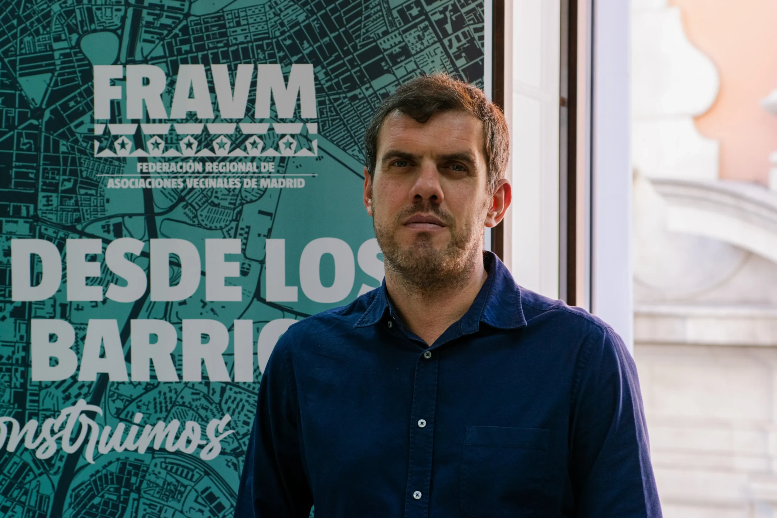 Jorge Nacarino, un hombre de pelo corto y castaño, con camisa azul, permanece de pie en un interior, delante de una pancarta verde azulado del Fravm con texto en blanco en español y un mapa gráfico de Madrid. La luz del sol entra por una ventana a la derecha, dejando entrever el vibrante ruido político de la ciudad.