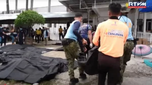 Varios equipos de emergencia y agentes de policía manipulan bolsas negras para cadáveres al aire libre cerca de un edificio en Filipinas, tras un terremoto, mientras los espectadores permanecen de pie al fondo; se ven lonas o más bolsas para cadáveres en el suelo.