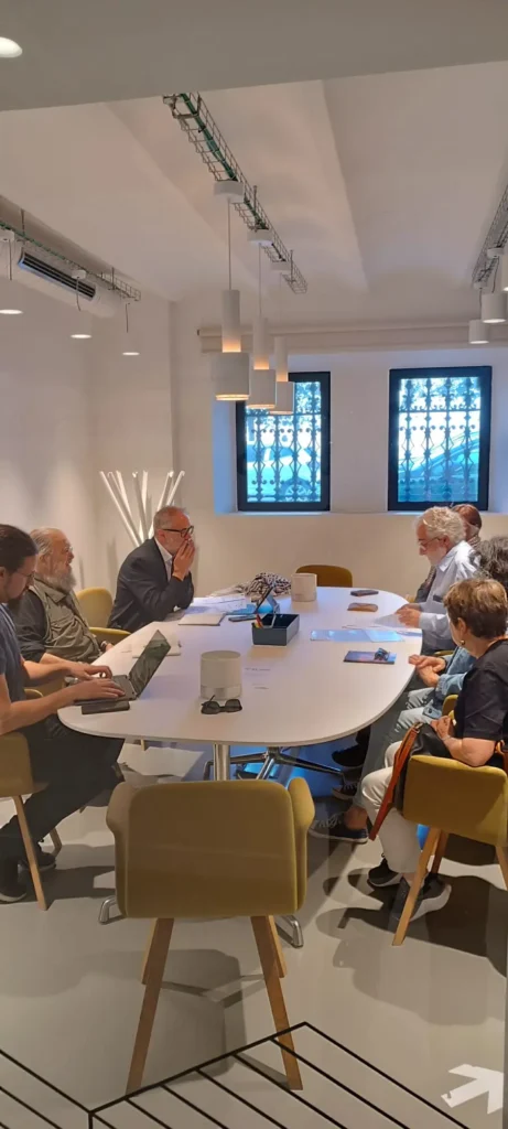 Siete personas se sientan alrededor de una mesa de conferencias blanca en una sala moderna, debatiendo temas como las inmatriculaciones y el papel de la Iglesia Católica, con ordenadores portátiles, cuadernos y papeles extendidos ante ellos.