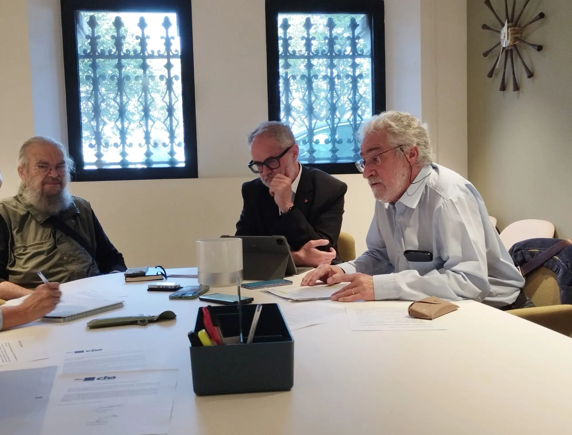 Tres hombres mayores se sientan a una mesa de conferencias en una sala con dos ventanas enrejadas, discutiendo inmatriculaciones que afectan a la Iglesia Católica. Sobre la mesa hay papeles, bolígrafos y una tableta mientras uno escribe, otro comprueba un dispositivo y el tercero mira a la cámara.