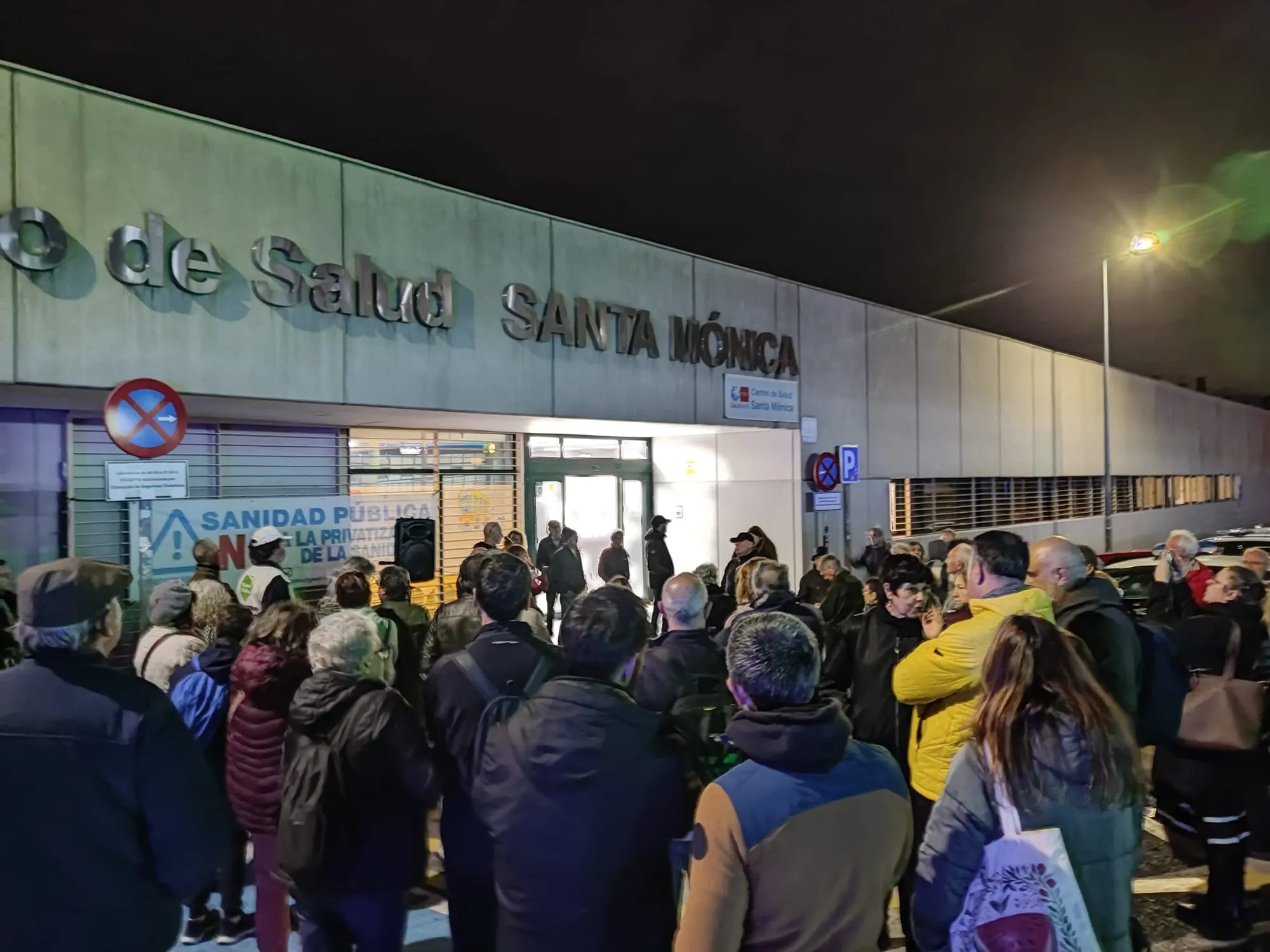 Un grupo se reúne por la noche ante el iluminado Centro de Salud Santa Mónica de Rivas, algunos con pancartas, en señal de manifestación pública en apoyo de la sanidad pública.