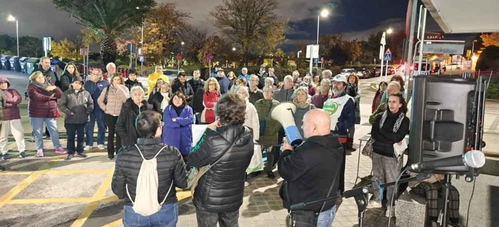 Un grupo de personas se reúne al aire libre en una zona pavimentada por la noche en Rivas, algunos con pancartas para apoyar la sanidad pública. Varias personas miran a la multitud, una de ellas hablando por megáfono. Las farolas iluminan los árboles y los coches del fondo.