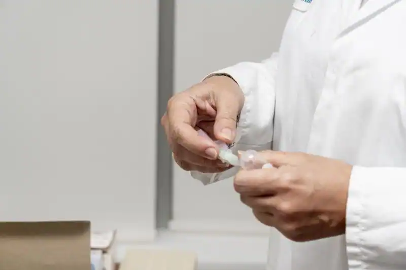 Una persona con bata blanca está abriendo un pequeño recipiente de plástico en un entorno clínico o de laboratorio, posiblemente preparándose para realizar pruebas de gripe o virus respiratorios, problemas de salud comunes en España. No se ve la cara de la persona.