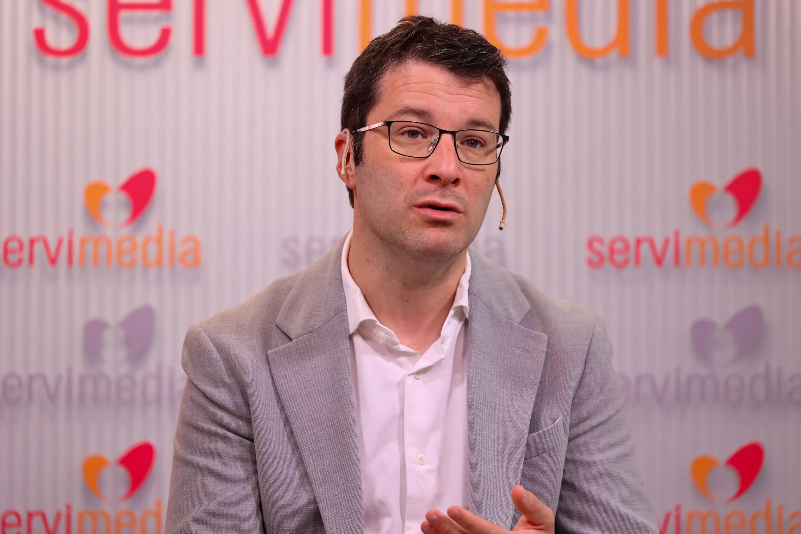 Un hombre con gafas, americana gris claro y camisa blanca habla mientras gesticula con la mano. Lleva un micrófono de diadema. El fondo muestra la palabra servimedia y referencias a la Plataforma de Infancia en texto rojo y naranja. Un hombre con gafas, americana gris claro y camisa blanca habla mientras gesticula con la mano. Lleva un micrófono de diadema. El fondo muestra la palabra servimedia y referencias a la Plataforma de Infancia en texto rojo y naranja.