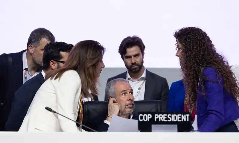 Un grupo de personas, entre ellas un hombre sentado detrás de un escritorio con la etiqueta COP PRESIDENT, participan en un debate sobre combustibles fósiles en una conferencia o reunión oficial. El fondo es claro y claro.