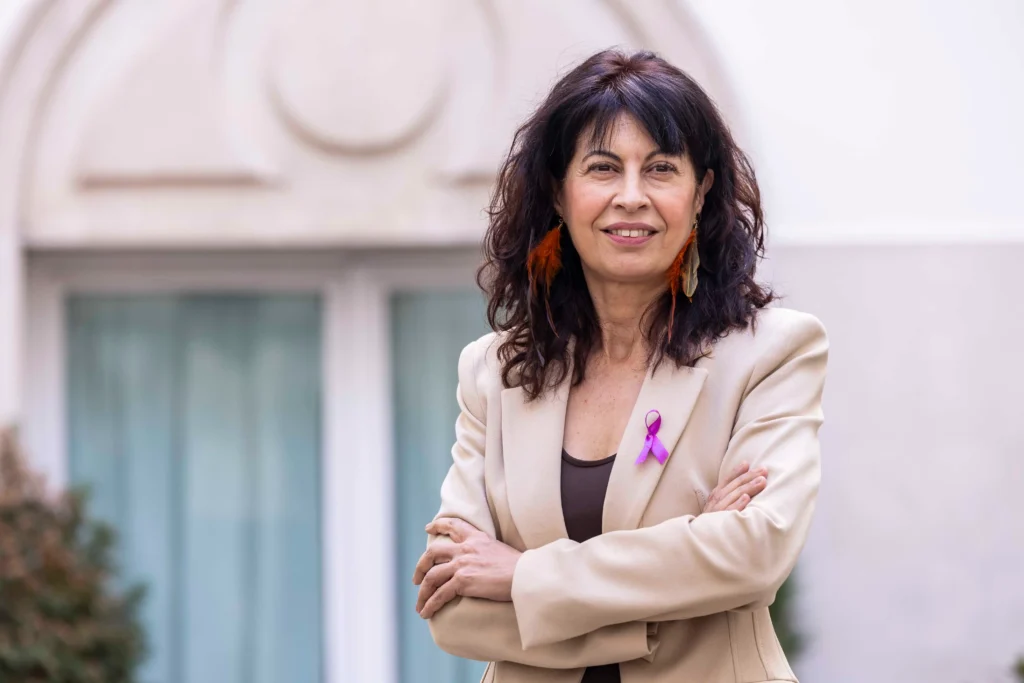 Una mujer con el pelo oscuro y ondulado está de pie al aire libre delante de un edificio. Lleva una americana beige con un lazo morado de concienciación, un pendiente naranja y los brazos cruzados mientras sonríe a la cámara.