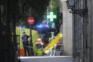 Dos socorristas se encuentran detrás de una cinta policial en una acera de la ciudad, uno de ellos sujetando una camilla amarilla. Al fondo se ve una ambulancia, agentes de policía y una cruz verde de farmacia, junto a un borrador automático aparcado.