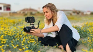 Una persona con el pelo rizado, vestida con una americana blanca y pantalones negros, se sienta entre flores silvestres amarillas mientras sostiene una cámara con un monitor conectado, captando la escena, tal vez contando historias de víctimas o destacando la desprotección judicial.