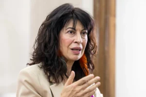 Una mujer con el pelo largo, ondulado y oscuro y una americana beige gesticula con la mano mientras habla de OnlyFans. Está en un interior y el fondo está desenfocado.