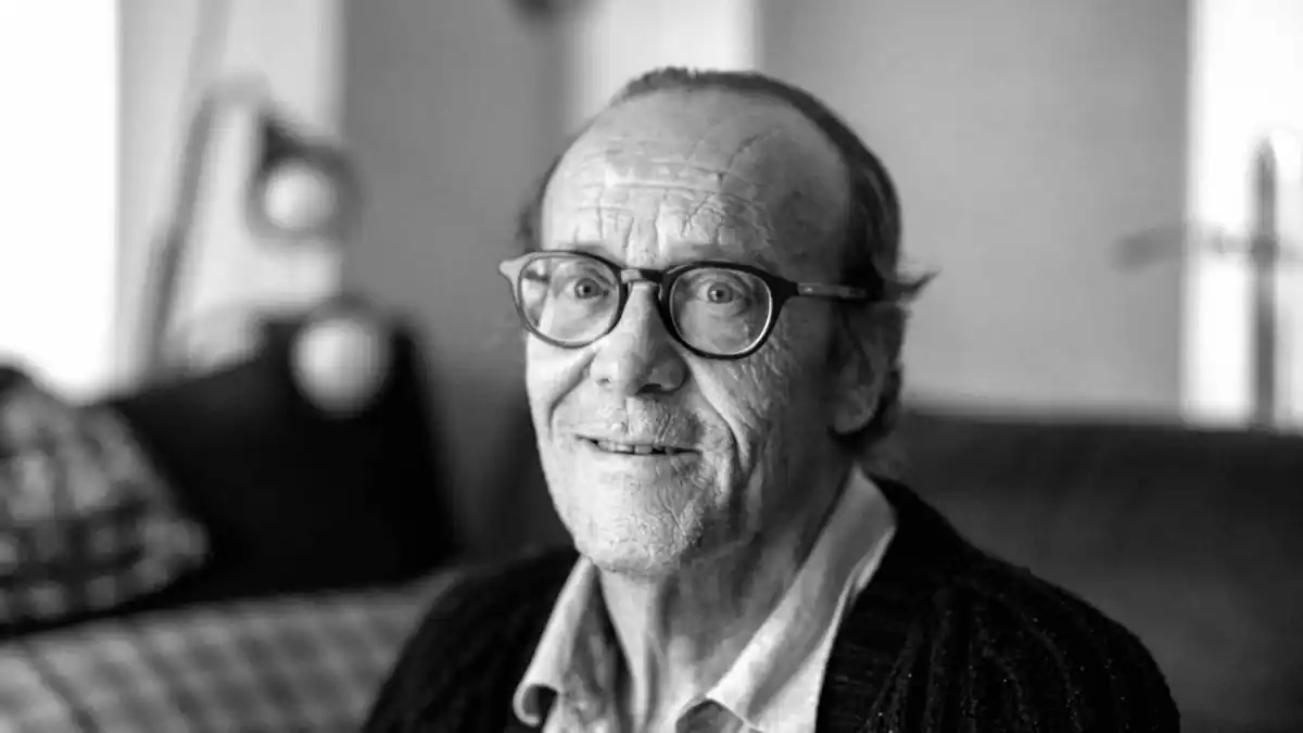 Un hombre mayor con gafas y el pelo ralo está sentado en el interior, mirando a la cámara y sonriendo ligeramente. La foto, en blanco y negro, tiene un fondo borroso con una lámpara y un cojín que evocan historias de la Puerta del Sol durante la época del expreso del franquismo.
