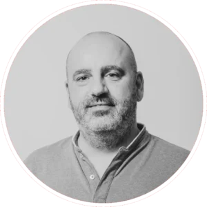 Un hombre de mediana edad, con la cabeza y la barba afeitadas y vestido con camisa de cuello, mira a la cámara con expresión neutra. La imagen en blanco y negro de Adolfo Calle está enmarcada por un borde circular.