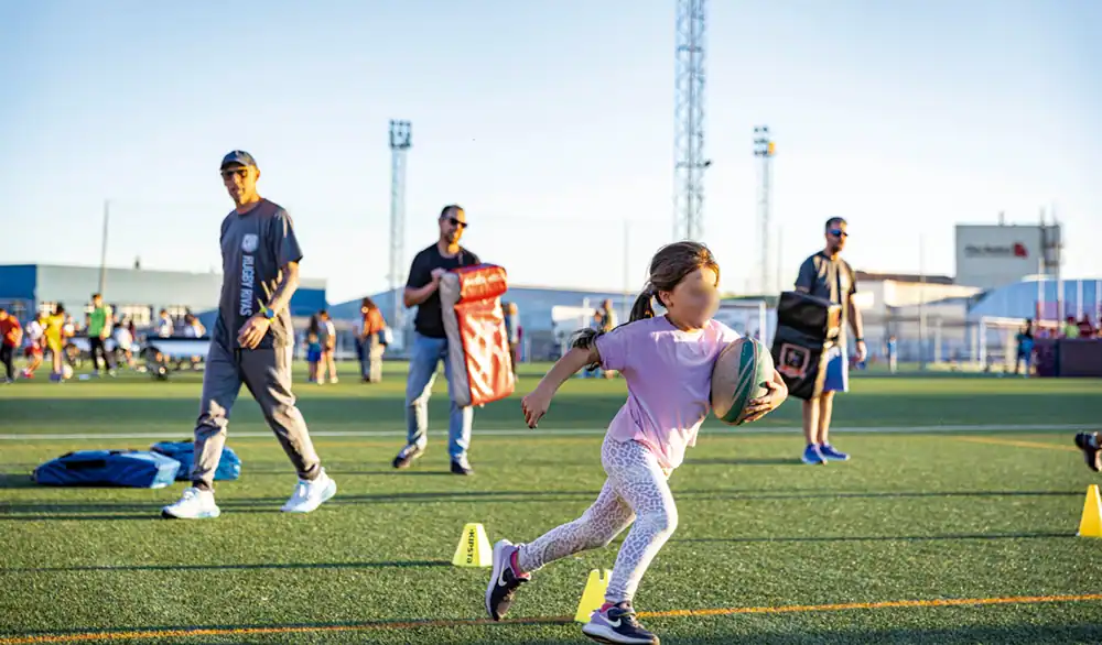 Un niño corre con un balón de rugby en un campo deportivo de Rivas, sorteando conos amarillos, mientras varios adultos y niños permanecen de pie al fondo, bajo un cielo despejado, durante la Semana Europea del Deporte. Un niño corre con un balón de rugby en un campo deportivo de Rivas, sorteando conos amarillos, mientras varios adultos y niños permanecen de pie al fondo, bajo un cielo despejado, durante la Semana Europea del Deporte.