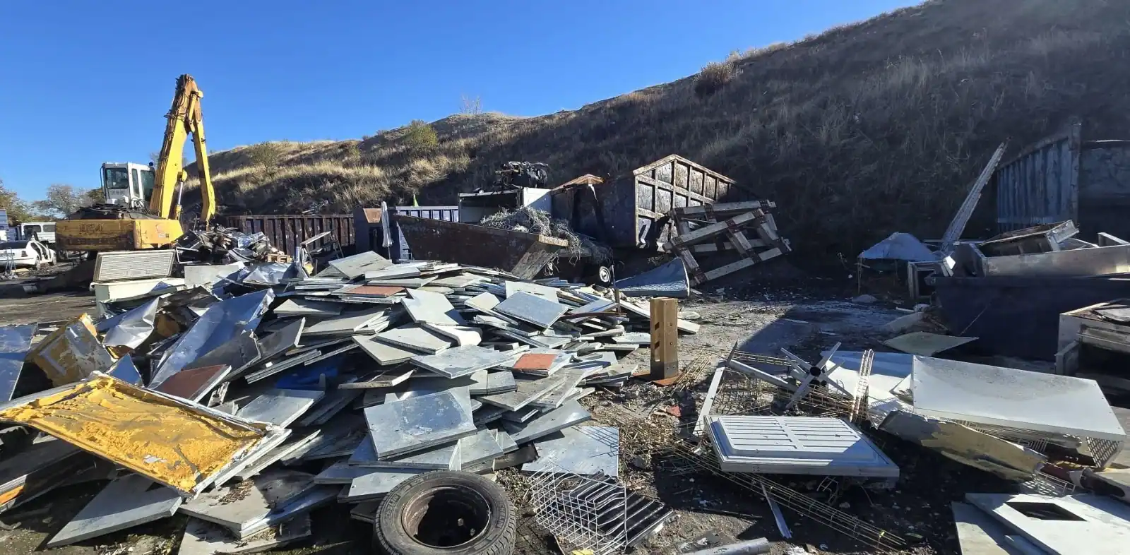Un montón de escombros metálicos, electrodomésticos y materiales de desecho se encuentra en un depósito de chatarra al aire libre cerca de una colina de tierra junto a la Cañada. Una excavadora amarilla está aparcada a la izquierda, rodeada de neumáticos y objetos rotos, vestigios de la industria ilegal, bajo un cielo despejado.