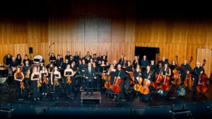 Una orquesta completa, compuesta por músicos de cuerda, viento-metal, viento-madera y percusión, posa en el escenario vestida de concierto con sus instrumentos, lista para interpretar música de cuerda o selecciones de ópera y zarzuela. El escenario presenta paneles de madera al fondo.