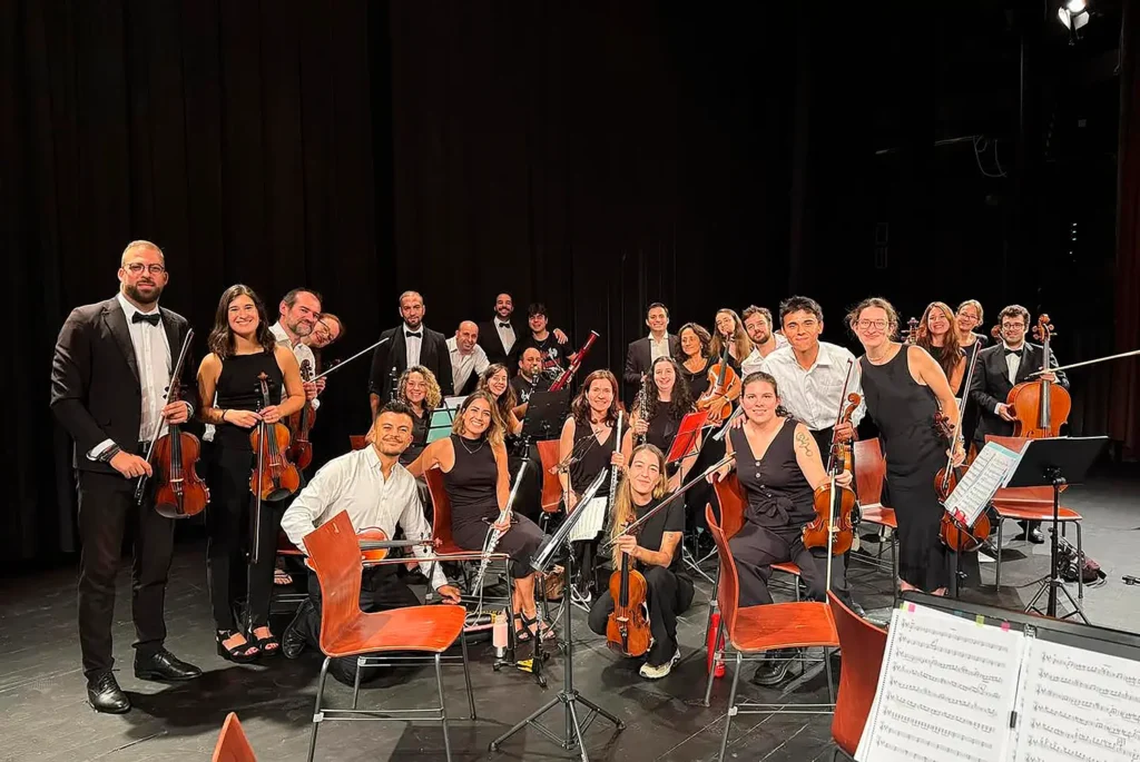 Un grupo de músicos vestidos de concierto posan en el escenario con sus instrumentos de cuerda y viento, listos para interpretar música de cuerda, rodeados de sillas vacías y atriles sobre un fondo oscuro.