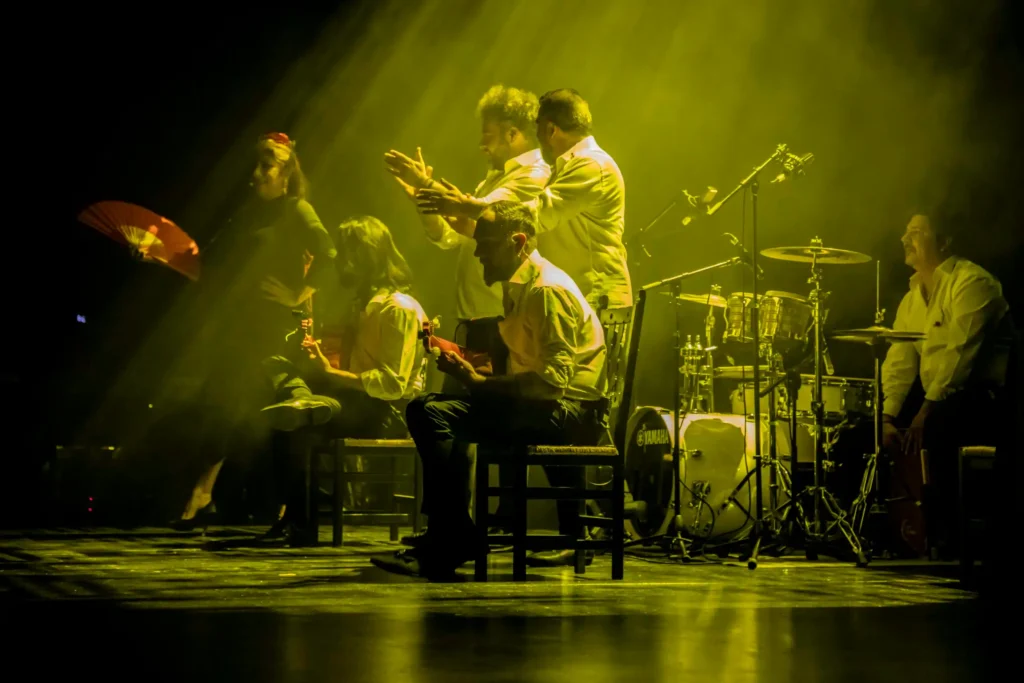Un grupo de seis artistas de Rivas Flamenca en el escenario bajo una iluminación amarilla; cuatro músicos sentados tocan la guitarra, una persona da palmas y una bailaora está de pie a un lado sujetando un abanico rojo. Al fondo, tambores y micrófonos.