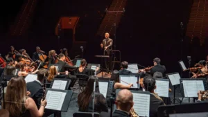 Un director de orquesta, de pie sobre un podio, dirige una orquesta de músicos que tocan varios instrumentos en el escenario, con partituras en atriles y filas de asientos vacíos visibles en el fondo poco iluminado.