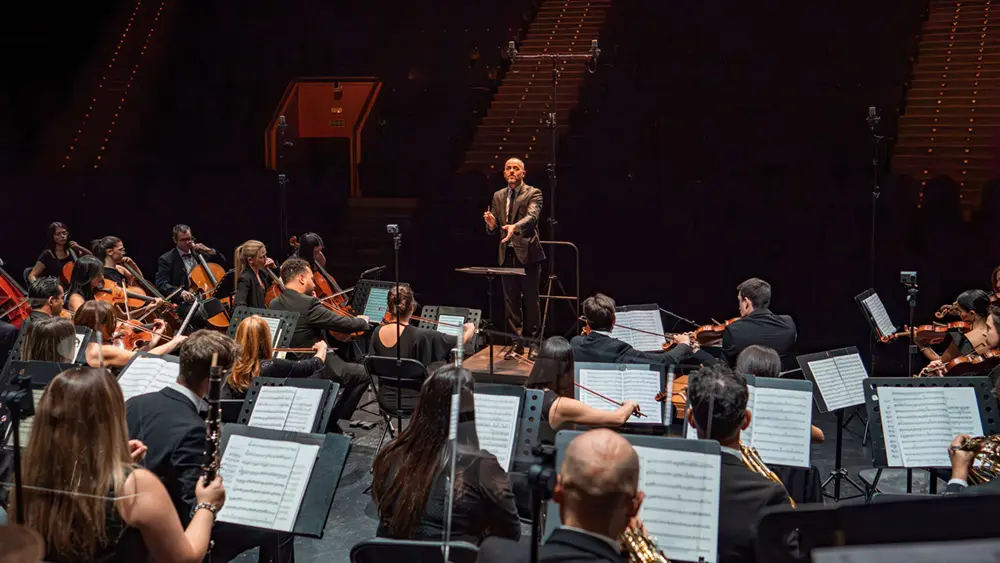 Un director de orquesta, de pie sobre un podio, dirige una orquesta de músicos que tocan varios instrumentos en el escenario, con partituras en atriles y filas de asientos vacíos visibles en el fondo poco iluminado.