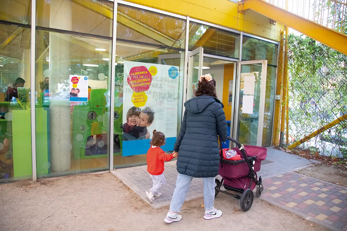 Una mujer con un cochecito y un niño pequeño entra en un edificio con grandes ventanas y puertas de cristal. La colorida señalización sugiere que se trata de una guardería o un centro comunitario, posiblemente con servicios como Borrador automático para familias.
