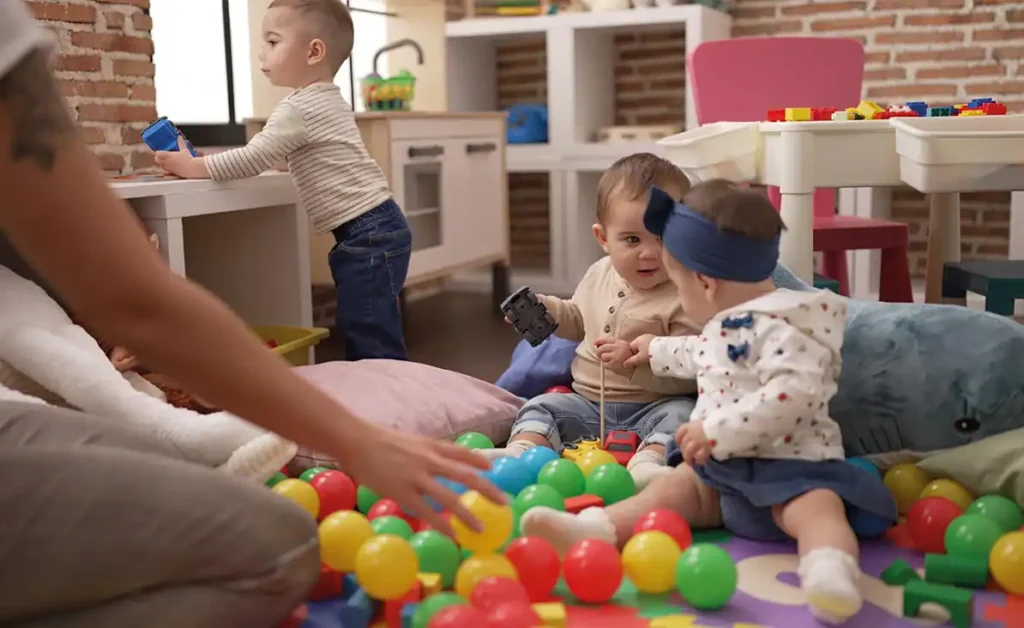 Tres niños pequeños juegan con pelotas y juguetes de plástico de colores en el suelo de una sala de juegos, explorando talleres creativos. Un niño está de pie junto a una cocina de juegos en el fondo mientras el brazo de un adulto se extiende hacia los niños desde el lado izquierdo.