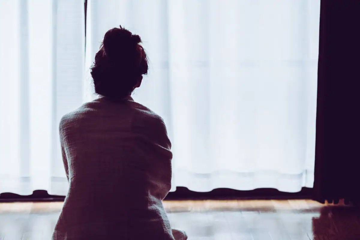Una mujer con el pelo recogido en un moño está sentada en el suelo, envuelta en una manta, frente a una ventana con cortinas blancas. La escena, suavemente iluminada, evoca la contemplación silenciosa, aludiendo a las luchas silenciosas a las que se enfrentan muchas mujeres en pareja en medio de la violencia machista. Una mujer con el pelo recogido en un moño está sentada en el suelo, envuelta en una manta, frente a una ventana con cortinas blancas. La escena, suavemente iluminada, evoca la contemplación silenciosa, aludiendo a las luchas silenciosas a las que se enfrentan muchas mujeres en pareja en medio de la violencia machista.