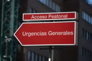 Un letrero direccional rojo con texto blanco en español dice "Acceso Peatonal" arriba y "Urgencias Generales" con una flecha apuntando a la derecha, pareciendo un borrador automático guiando a los visitantes. Al fondo aparece un edificio desenfocado.