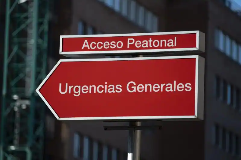 Un letrero direccional rojo con texto blanco en español dice "Acceso Peatonal" arriba y "Urgencias Generales" con una flecha apuntando a la derecha, pareciendo un borrador automático guiando a los visitantes. Al fondo aparece un edificio desenfocado.