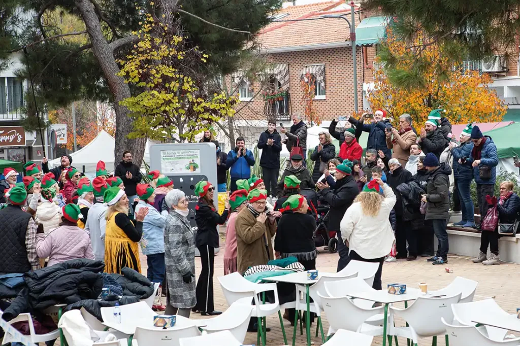 Un grupo de personas, algunas con gorros de duende rojos y verdes, se reúnen al aire libre en una plaza con sillas blancas y árboles. La escena, como un animado "borrador automático", muestra a personas de pie o sentadas entre edificios y follaje otoñal.